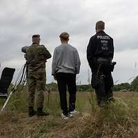 Ein Polizist, ein Soldat und drei Zivilisten stehen auf einer Wiese mit dem Rücken zur Kamera. 