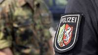 Zusammenarbeit Das Wappen der Landespolizei Nordrhein-Westfalen. Im Hintergrund steht ein Soldat in Uniform.