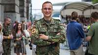 Ein Soldat steht auf einem Weg und lächelt in die Kamera, im Hintergrund sind Menschen zu sehen
