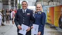 Ein Feuerwehrmann und eine Soldatin stehen auf einem Weg und halten Urkunden in den Händen