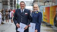 Eine Feuerwehrmann und eine Soldaten stehen auf einem Weg und halten jeweils eine Urkunde in den Händen