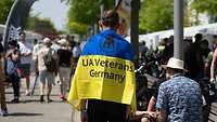 Ein Mann mit einer Flagge in den farben der Ukraine mit der Aufschrift "UA Veterans Germany" steht auf einem Weg 