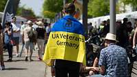 Ein Mann hat eine Ukraine-Flagge mit der Aufschrift "UA Veterans Germany" um die Schulter gelegt