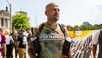 Ein Mann mit einem T-Shirt mit der Aufschrift "Sportkamerad" steht vor dem Bundestag