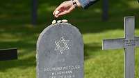 Ein Grabstein mit einem jüdischen Stern und einer Hand, die von oben Steine auf die Grabstele legt
