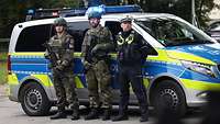 Teaserbild Zwei Soldaten stehen mit einem Polizisten vor einem Polizeiauto.