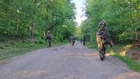 Sechs als Schützenreihe aufgestellte Soldaten patrouillieren auf einem sandigen Waldweg.