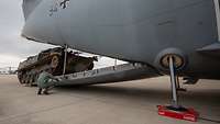Am Heck eines grauen Flugzeugs befährt ein Panzer die Heckrampe. Daneben steht ein Soldat.