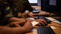 Drei Soldaten sitzen an einem Tisch vor Computern. Einer schreibt etwas auf ein Blatt und einer tippt auf der Tastatur.