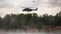 Zwei Hubschrauber fliegen über Wasser. Sie transportieren unter sich einen Behälter. 