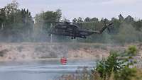 Ein Hubschrauber fliegt über Wasser. Er transportiert unter sich einen Behälter.
