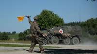 Ein Soldat schwenkt eine gelbe Flagge. Im Hintergrund fährt ein Militfärfahrzeug.