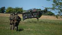 Eine Drohne landet auf einer Wiese. Zwei Soldaten tragen einen Verwundeten zur Drohne. 