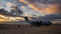 Ein großes graues Transportflugzeug steht auf einer weiten Betonfläche, daneben Soldaten.