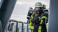 Zwei Feuerwehrmänner stehen bei der Übung Northern Coasts 2024 in voller Montur auf einem Schiff.
