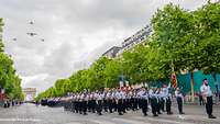 Soldatinnen und Soldaten stehen in Formation auf einer Avenue, am Himmel sind vier Flugzeuge. 