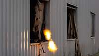 Ein Maschinengewehr schießt mit hellem Feuer aus der Fensterluke einer Blechbaracke.