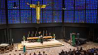 Eine blaue Wand mit einem goldenen Jesus, im Vordergrund ein Altar und einer Frau, die Andacht hält.