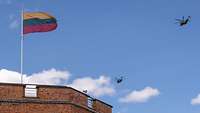 Auf einem Turm weht die litauische Flagge und zwei Hubschrauber fliegen am Himmel