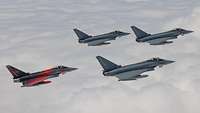 Vier Kampfflugzeuge, einer davon mit Sonderfolierung, fliegen in Formation über den Wolken.