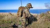 Ein Soldat hockt an einem Küstenabschnitt im Gelände. Rechts im Bild steigt Rauch auf.