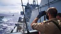Ein uniformierter Soldat schaut durch ein Fernglas von einem Schiff aus aufs Meer hinaus.