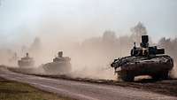 Mehrere Panzer fahren in eine Staubwolke gehüllt hintereinander durchs Gelände.