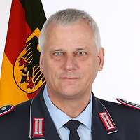 Portrait von Oberst i.G. Dr.-Ing. Wolfgang Smeets mit einer Truppenfahne im Hintergrund.