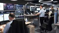 Soldaten und Zivilisten stehen und sitzen vor Computern in einem Lagezentrum.