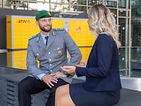 Ein Soldat und eine Frau sitzen vor einer DHL-Packstation und sprechen miteinander