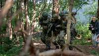 Zwei Soldaten stützen einen Soldaten, während sie durch ein Waldstück laufen, dahinter filmt ein Mann
