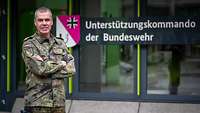 Ein Soldat in Uniform steht vor einem Gebäude mit Wappen und Schriftzug