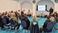 Menschen sitzen auf Stühlen und schauen zu einem Imam