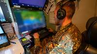 Ein Soldat sitzt mit Kopfhören vor einem Computer mit zwei übereinander angeordneten Bildschirmen.