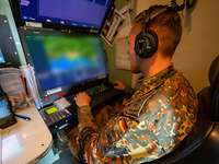 Ein Soldat sitzt mit Kopfhören vor einem Computer mit zwei übereinander angeordneten Bildschirmen.