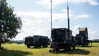 Drei militärische Lastwagen, einer mit einem aufgerichteten Radar, stehen auf einer Wiese.