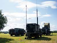 Drei militärische Lastwagen, einer mit einem aufgerichteten Radar, stehen auf einer Wiese.