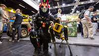Ein Mann in einem Wolfskostüm posiert mit einem gelben Roboterhund der Firma Boston Dynamics