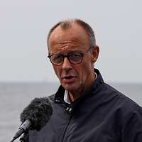 Ein Mann spricht in ein Mikrofon, im Hintergrund sind Schiffe der Deutschen Marine in See.