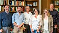 Zwei Frauen und vier Männer stehen in einer Bibliothek und schauen in die Kamera