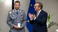 Ein Soldat hält eine Medaille, daneben steht Verteidigungsminister Boris Pistorius und applaudiert.