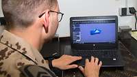 Blick über die Schulter eines Soldaten, der an einem Laptop sitzt. Auf dem Bildschirm eine blaue Form.