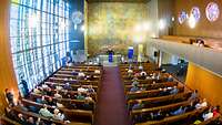 Ein Kirchsaal mit Menschen in den Bankreihen, ein Bischof spricht an einem Pult.