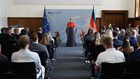 Mehrere Zivilistinnen und Zivilisten sitzen mit Blickrichtung zur Bühne in einem Saal. Am Pult spricht eine Frau zum Publikum.