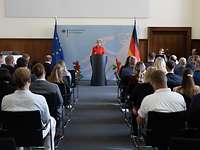 Mehrere Zivilistinnen und Zivilisten sitzen mit Blickrichtung zur Bühne in einem Saal. Am Pult spricht eine Frau zum Publikum.