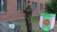 Eine Soldat posiert vor einem Gebäude für ein Foto, daneben ein Aufsteller mit dem Verbandsabzeichen