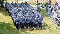 Soldaten in Dienstanzug marschieren in Formation auf eine Wiese an Schaulustigen vorbei