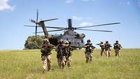 Hubschrauber steht auf grüner Wiese; davor laufen Soldaten mit Gewehren im Anschlag