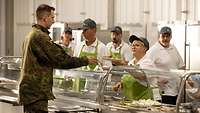 Ein Soldat steht vor einem Theke und bekommt von einer Frau einen Teller mit Essen gereicht