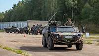 Ein Militärfahrzeug fährt vor einer Waldkante. Im Hintergrund weitere Fahrzeuge und Garagen.